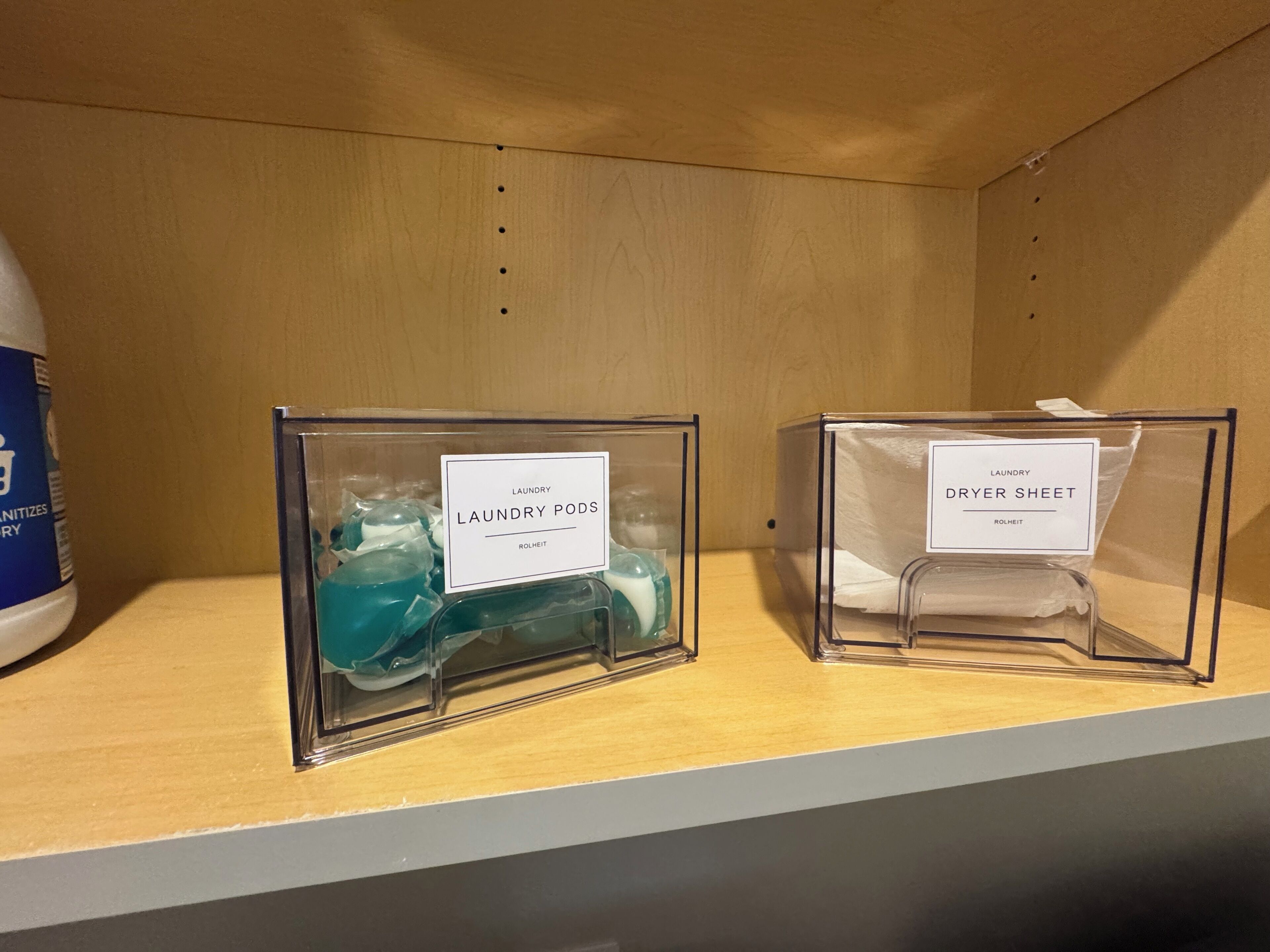 Twee transparante plastic opbergbakken op een plank, waarvan er een is gelabeld 'Laundry Pods' en de andere 'Dryer Sheet'.