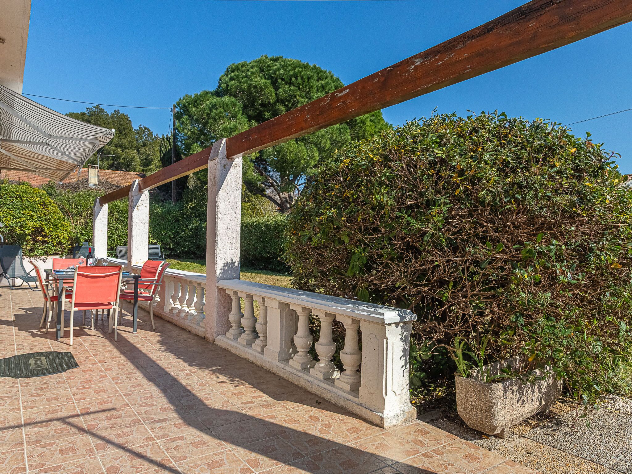 Un patio al aire libre con piso de baldosas, una pérgola de madera, una zona de estar con sillas rojas y una planta grande en maceta.