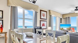 A dining area with a glass table and chairs, a view of the ocean through large windows, and a ceiling fan.