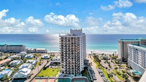 A high-rise hotel with a view of the ocean, surrounded by smaller buildings and a parking area.