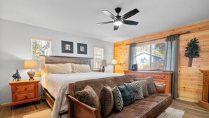 Une chambre à coucher comprenant un lit, une tête de lit en bois, un ventilateur de plafond, une table de chevet, une commode et une fenêtre avec des rideaux.