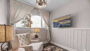 A cozy room with a white upholstered armchair, a small side table with a lamp, a fluffy rug, and a painting of a sunset on the wall. - Bunny's Ranch - Sleeps 12, Private Ranch in Big Bear, Pool Table, Ping Pong, Foosball, BBQ (Big Bear City)