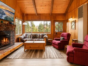 L’intérieur chaleureux d’un chalet avec une cheminée, un canapé, deux fauteuils et de grandes fenêtres donnant sur le paysage extérieur.