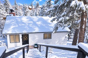 A white cabin with a black door and windows, surrounded by snow-covered trees and a wooden deck.