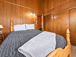 A room with wooden panel walls, a bed with a grey patterned bedspread, a white pillow, and a grey blanket. There is a wooden nightstand with a lamp and books.
