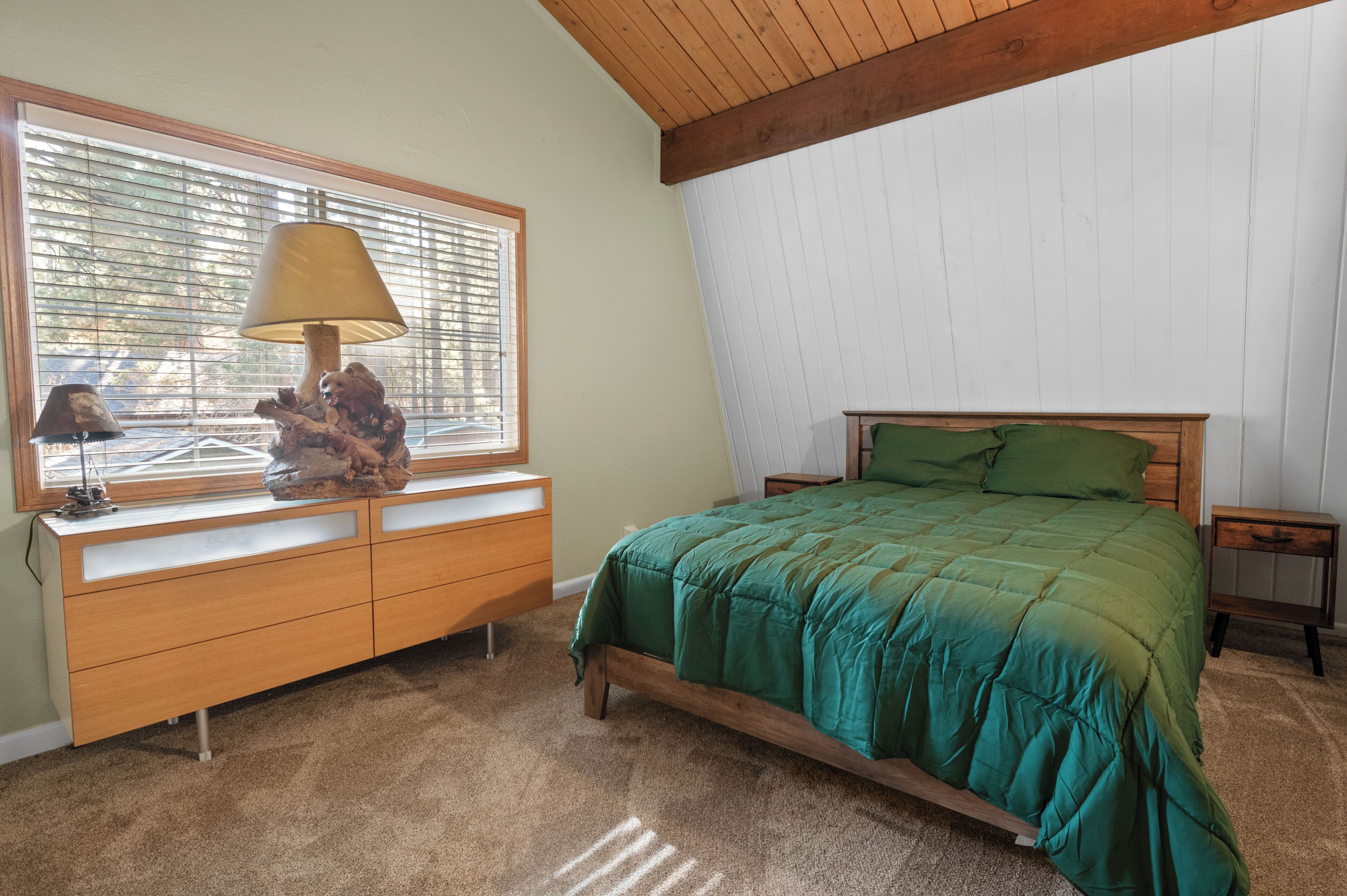 A bedroom with a wooden bed, a green bedspread, a wooden dresser, a lamp, and a window with blinds.