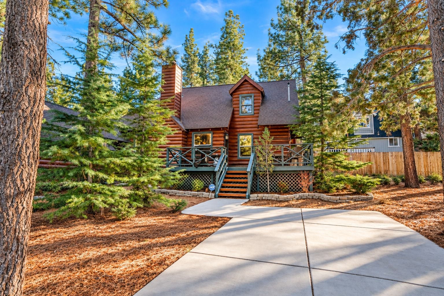 A log cabin with a wooden deck, a chimney, and a fenced yard surrounded by pine trees.
