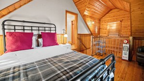 A cozy bedroom with a sloped ceiling, a bed with a plaid blanket, two bright red pillows, and a nightstand with a lamp.