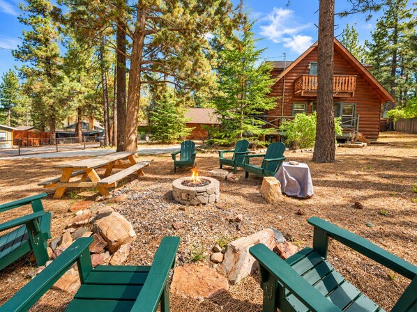 Una cabaña rústica con un hogar, una mesa de picnic y sillas Adirondack en un entorno boscoso.
