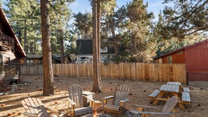 A backyard with a fire pit surrounded by Adirondack chairs, a wooden fence, and a cabin in the background.