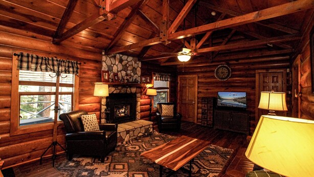 A log cabin interior with a fireplace, leather chairs, a television, and a patterned rug.