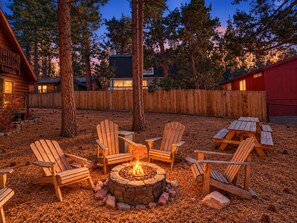 Une terrasse en bois, dotée de chaises Adirondack et d’un foyer en pierre, le tout entouré d’arbres.