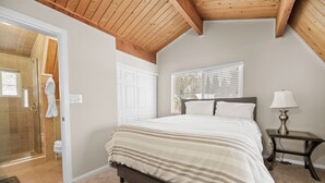 Une chambre avec un grand lit, une table de chevet avec une lampe, une fenêtre avec des stores et une porte donnant sur une salle de bain.