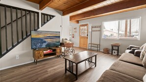 Un salon spacieux avec un escalier en bois, une grande fenêtre avec des stores, un canapé confortable et un téléviseur posé sur un meuble en bois.