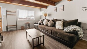 Un salon chaleureux avec un canapé en cuir marron, une table basse en bois et un banc. Il y a deux fenêtres équipées de stores, une lampe et une étagère fixée au mur.