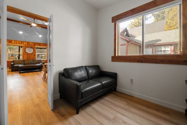 A room with a black leather sofa, wooden flooring, a large window with a view of a house, and a ceiling fan.
