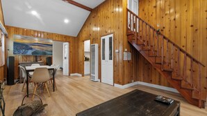 A spacious living area with a wooden staircase, a dining table with chairs, and a large painting on the wall. - Big Bear Cottage - Walk to Lake, Apple TV, Fireplace, Deck (Big Bear Lake)