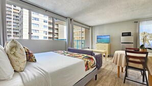 A hotel room with a bed, a desk, a chair, a TV, and a view of a building with balconies.