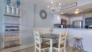A dining area with a round table and four chairs, a chandelier, a kitchen with white cabinets, and a sign that reads "Beach Life".