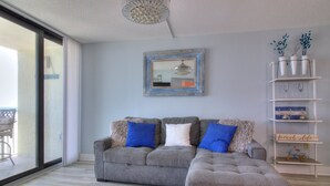 A living room with a grey sectional sofa, a small ottoman, a glass door leading to a balcony, a decorative mirror, and a shelving unit with decorative items.