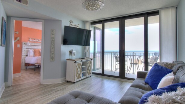 A spacious living room with a large gray sofa, a flat-screen TV mounted on the wall, and a balcony with a view of the beach.