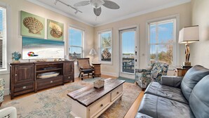 Un salon spacieux doté d’une grande fenêtre, d’un ventilateur de plafond, d’un meuble de rangement en bois foncé et d’un canapé en cuir gris.