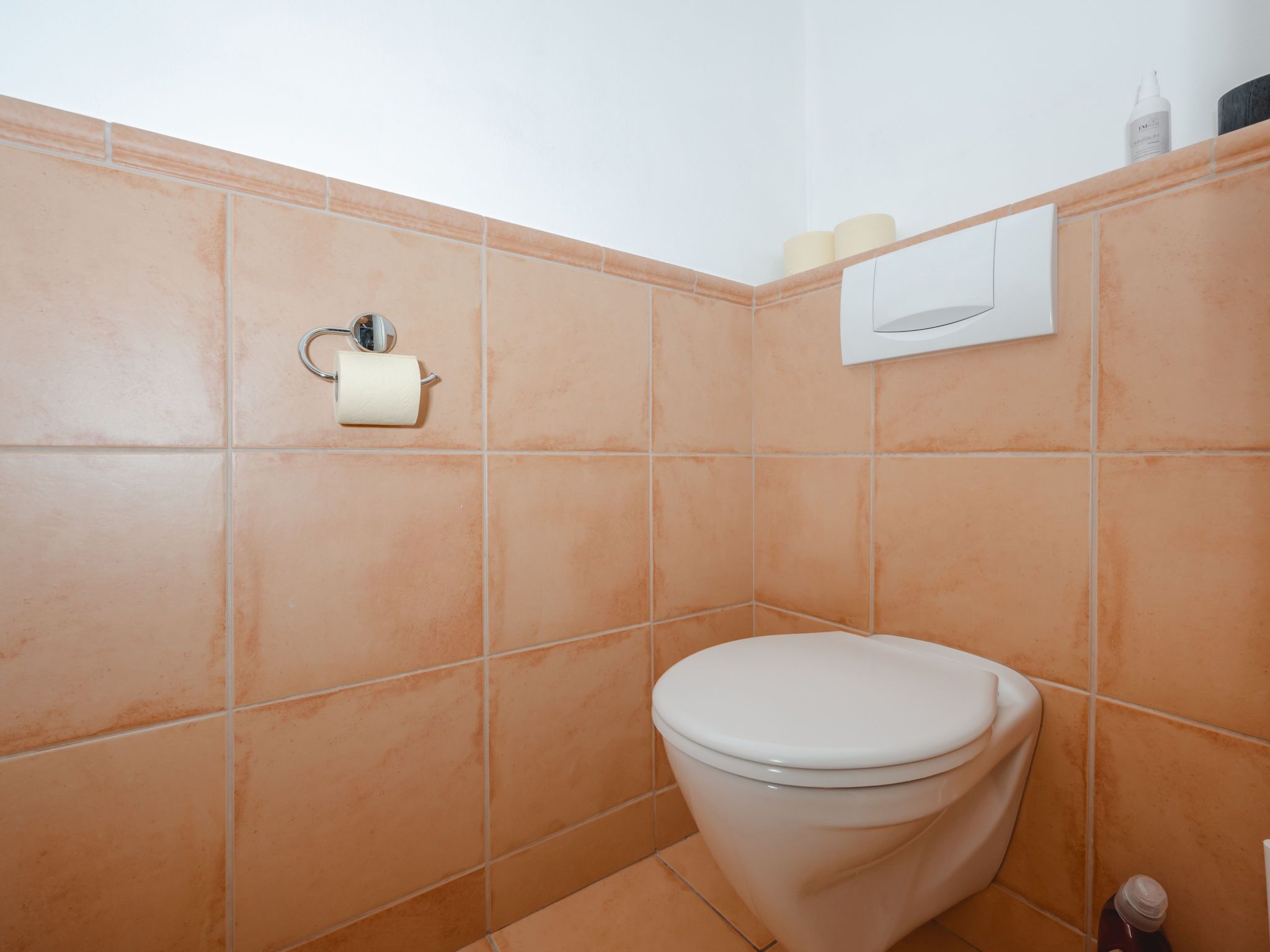 A bathroom with a white toilet, orange tiles, a toilet paper holder, and a hand dryer.