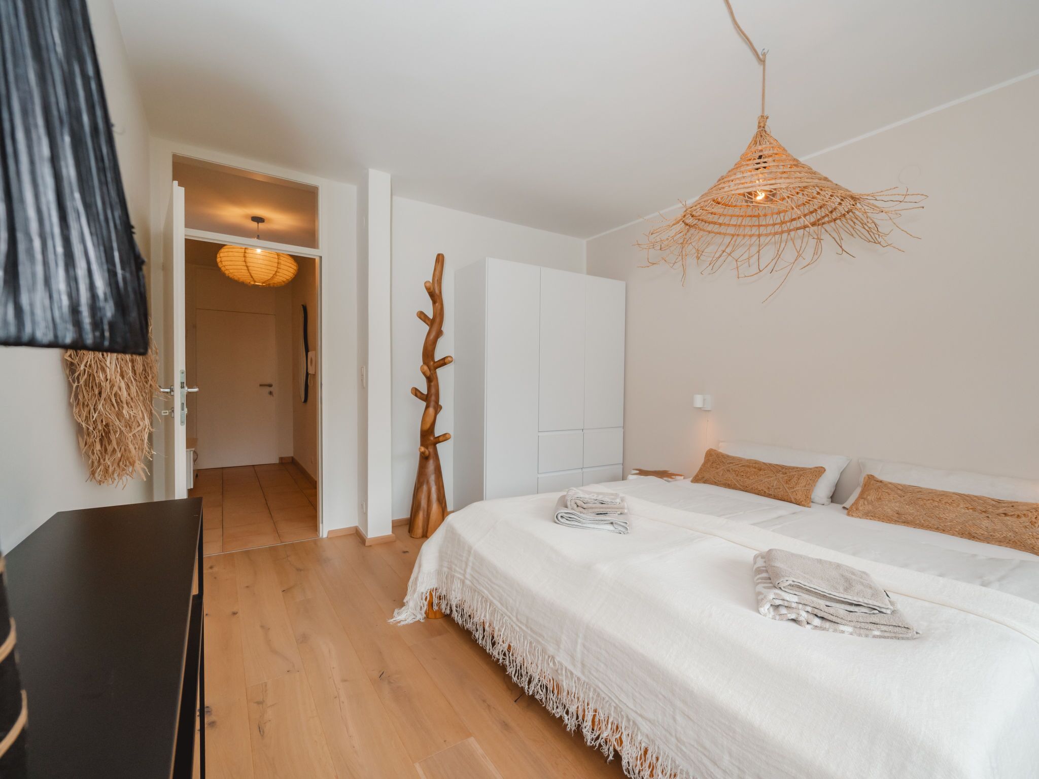 Camera da letto con un letto grande, un appendiabiti in legno, una lampada intrecciata e vista sul corridoio.