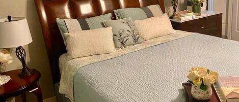 A neatly made bed with a light blue quilt, a wooden headboard, and multiple pillows. There is a small table with a lamp and a vase of flowers beside the bed. A framed picture hangs on the wall above the bed.