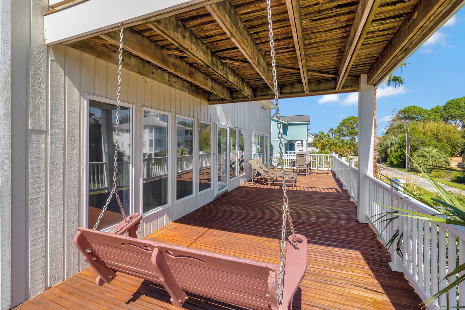A covered wooden deck with a swing set and a bench, overlooking a residential area with a house and palm trees.