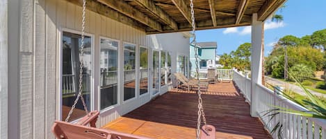 A covered wooden deck with a swing set and a bench, overlooking a residential area with a house and palm trees.