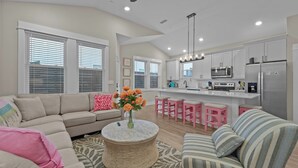 Una sala de estar moderna con un sofá, una mesa de centro y una cocina con armarios blancos y taburetes rosa.