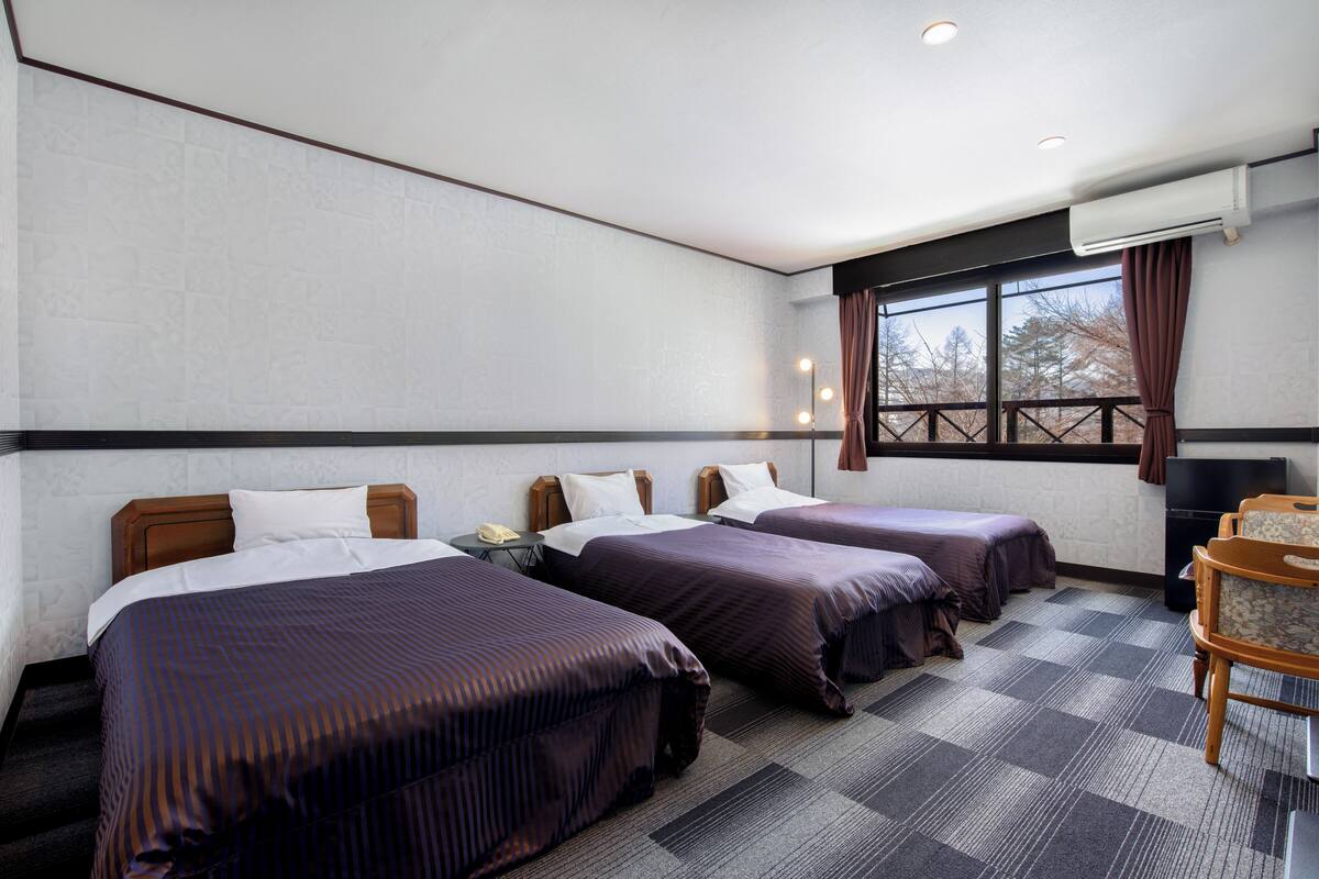 A hotel room with two beds, a desk, a chair, a window with curtains, and an air conditioning unit.