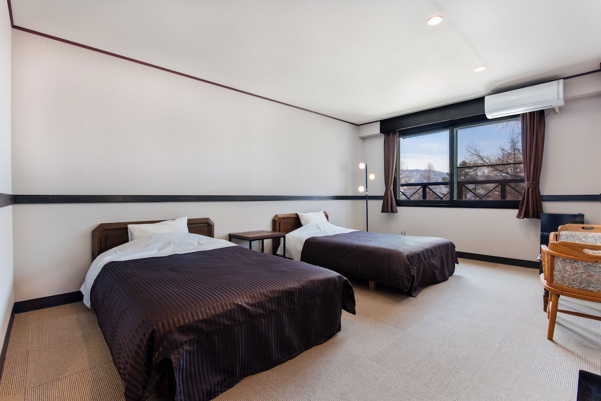 A hotel room with two beds, a desk, a chair, a window with a view, and an air conditioning unit.