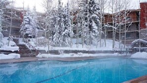 A swimming pool with a clear blue water surface, surrounded by snow-covered trees and a building with multiple balconies in the background.