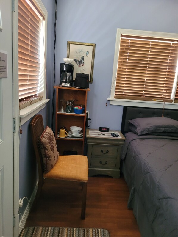A bedroom with a bed, a chair, a nightstand, a shelf with kitchen items, and a window with blinds. - The manner EN suite, marvelous 1 bedroom suite in moss Town exuma with AC, WiFi (Nassau bahamas)