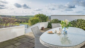 A round glass table with a wicker chair, a vase with flowers, and a plate with pastries on a rooftop terrace with a view of the countryside. - Ty Gwyn, Holiday Cottage Manorbier, Sleeps 8, 4 bedrooms,  bathrooms (Tenby)