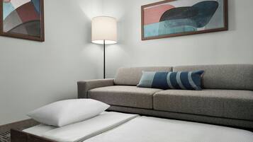 Une chambre moderne avec un canapé gris, un lit blanc doté d’une tête de lit en bois, une lampe sur pied et des œuvres d’art abstraites accrochées aux murs.