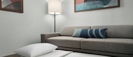 Una habitación moderna con un sofá gris, una cama blanca con cabecera de madera, una lámpara de pie y arte abstracto en las paredes.