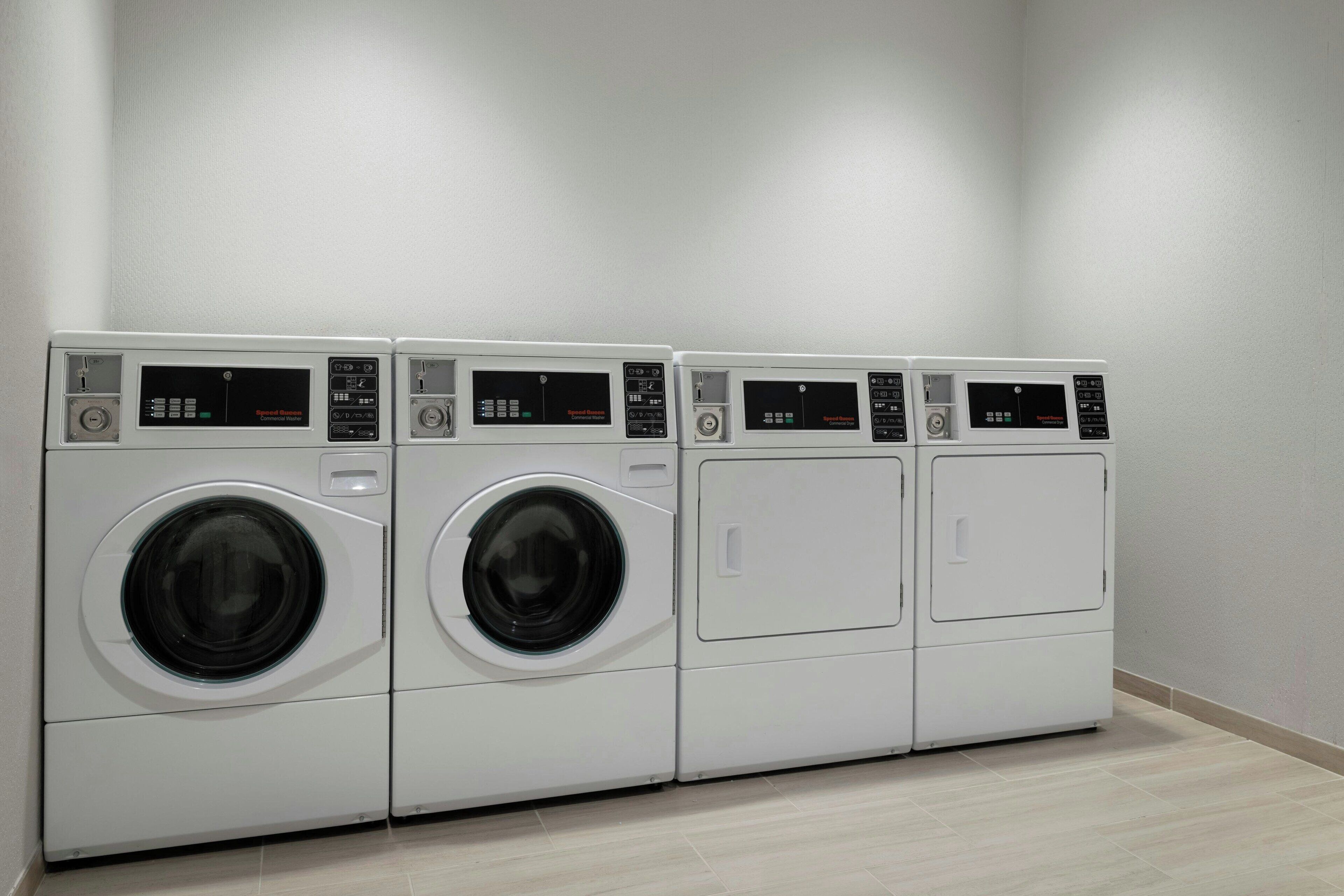 Een rij van vier witte wasmachines met voorlaadklep in een wasserij met een effen witte muur en een lichtgekleurde betegelde vloer.