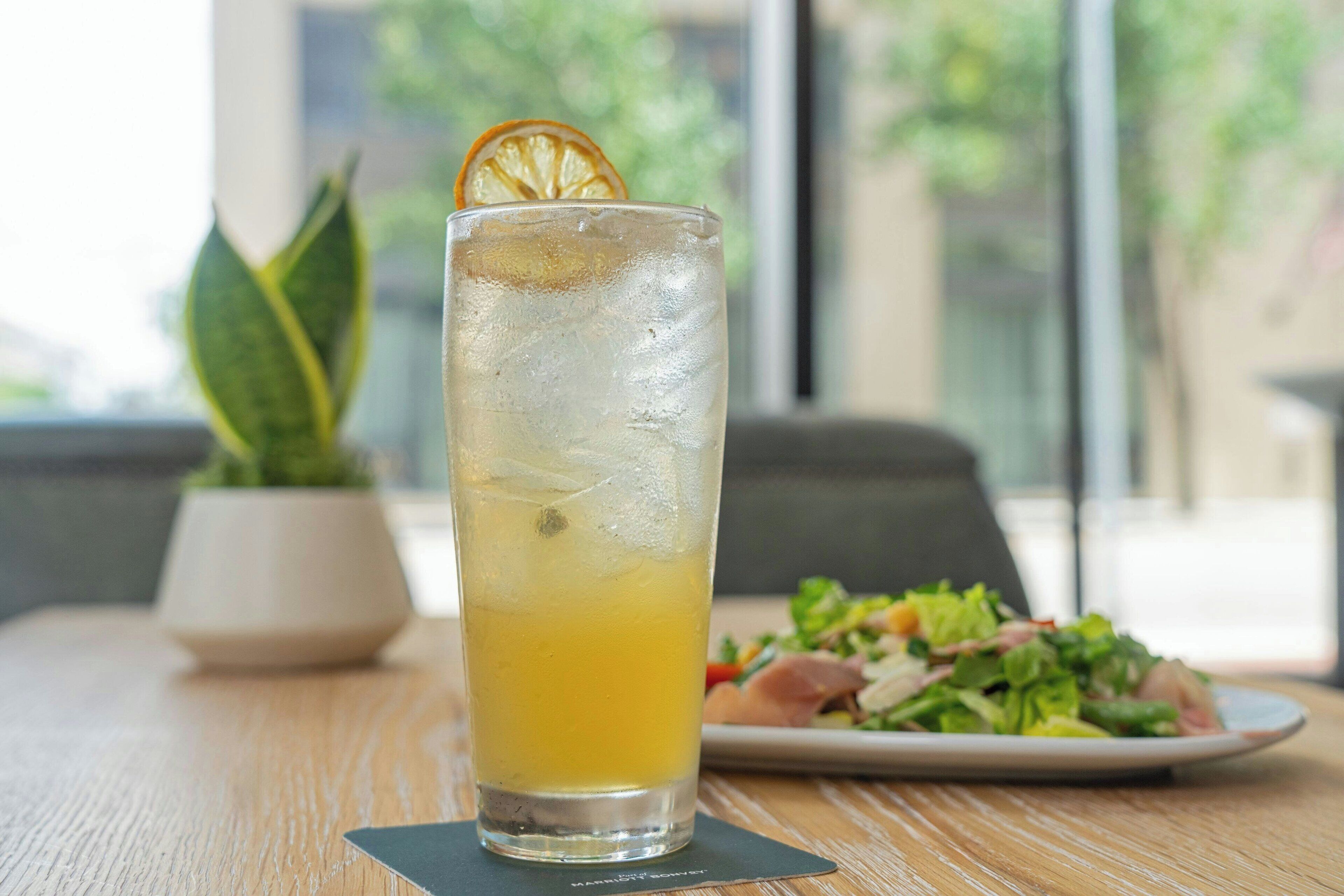 A tall glass of iced beverage with a slice of orange on the rim, placed on a wooden table with a plate of salad in the background.