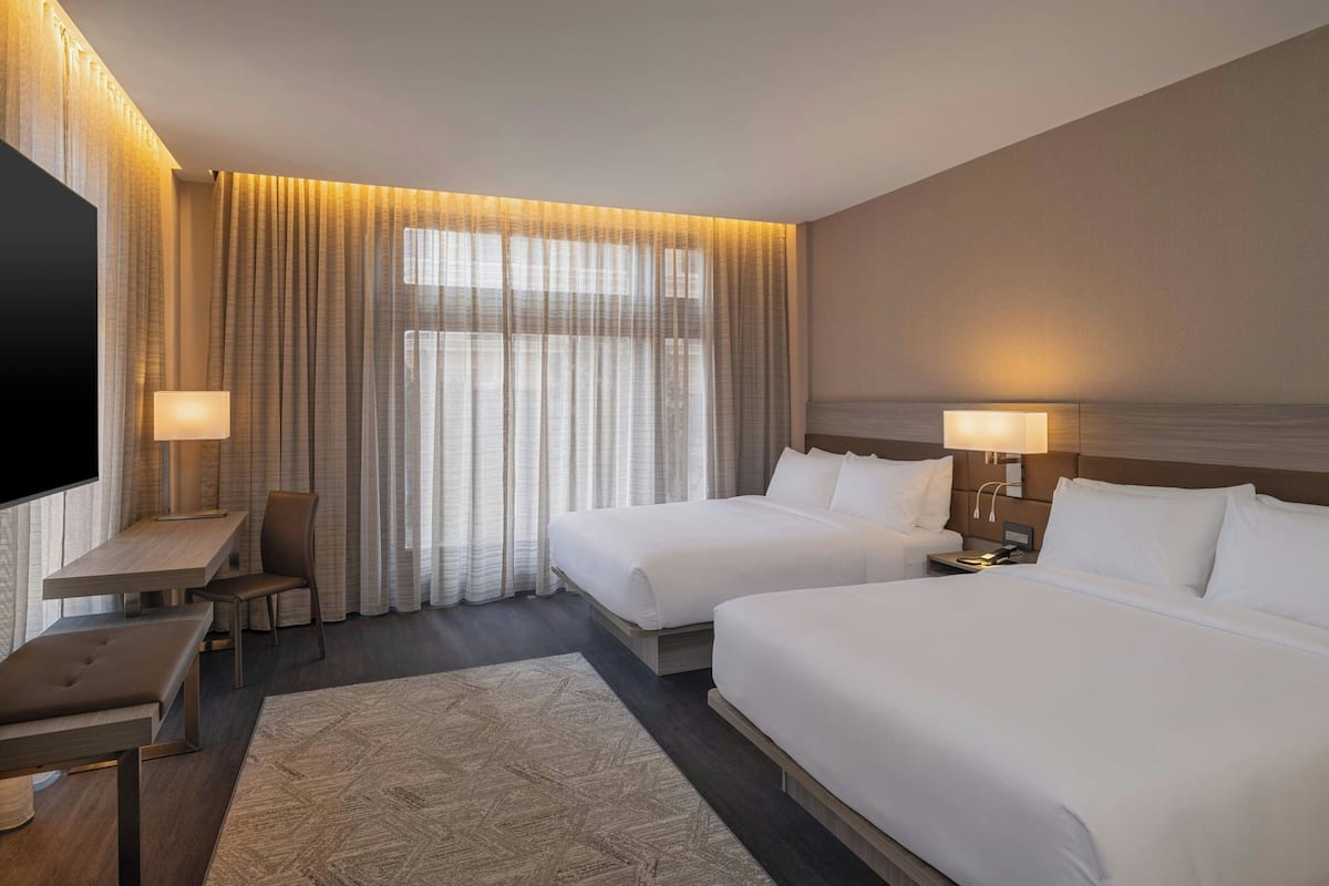 Una habitación de hotel con dos camas, un escritorio, una silla, un televisor y un ventanal grande con cortinas.