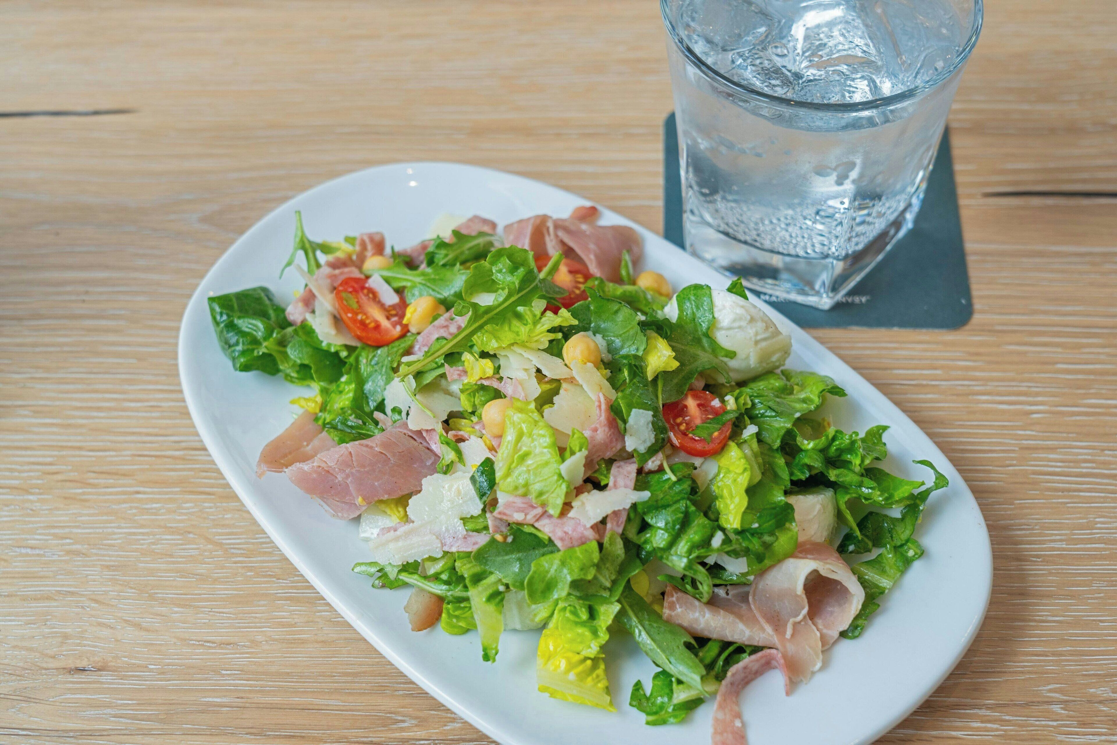 A plate of mixed greens salad with tomatoes, cheese, and ham on a wooden table.