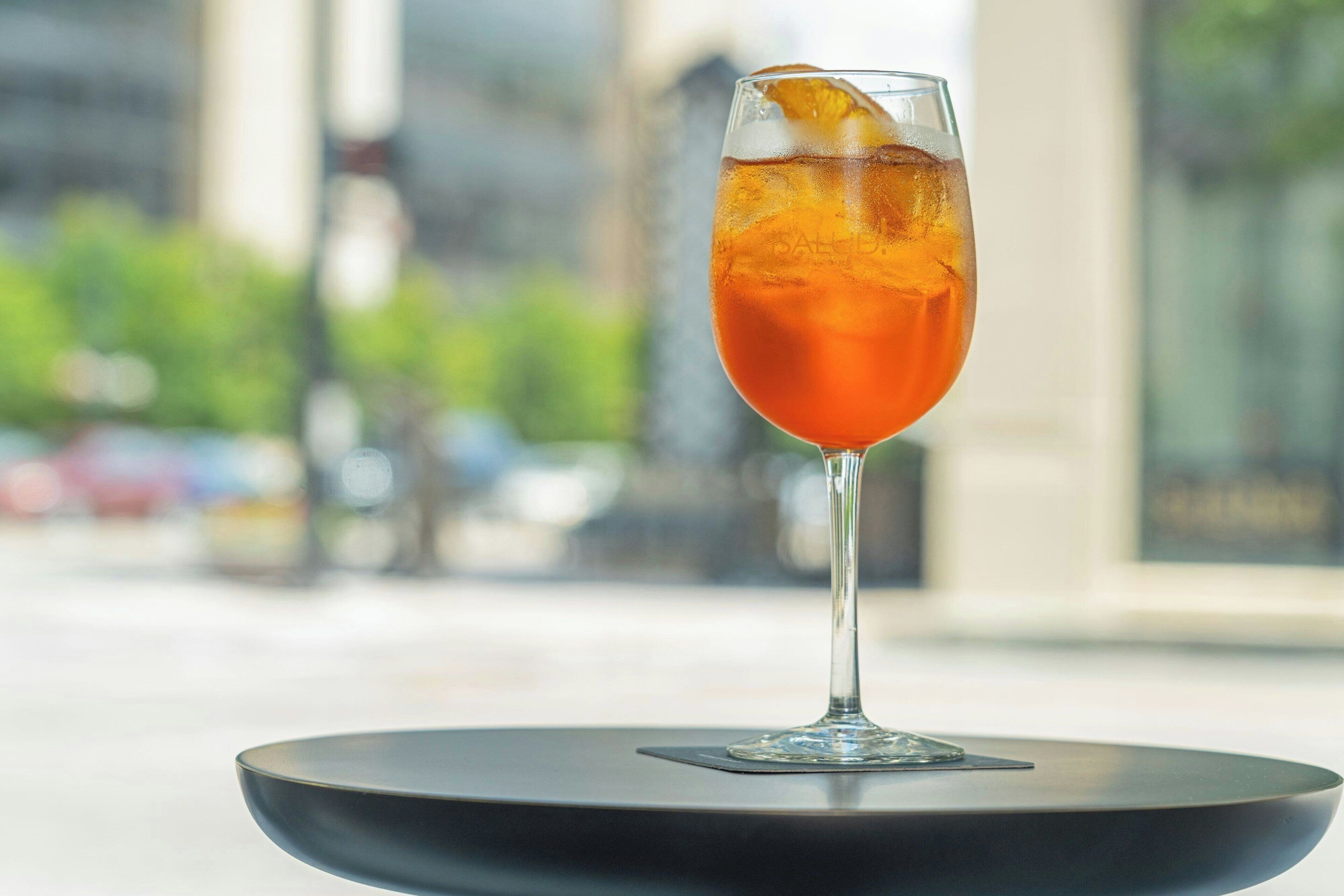 Un verre d’une boisson ambrée posé sur une table ronde noire, avec en arrière-plan une vue floue de la ville.
