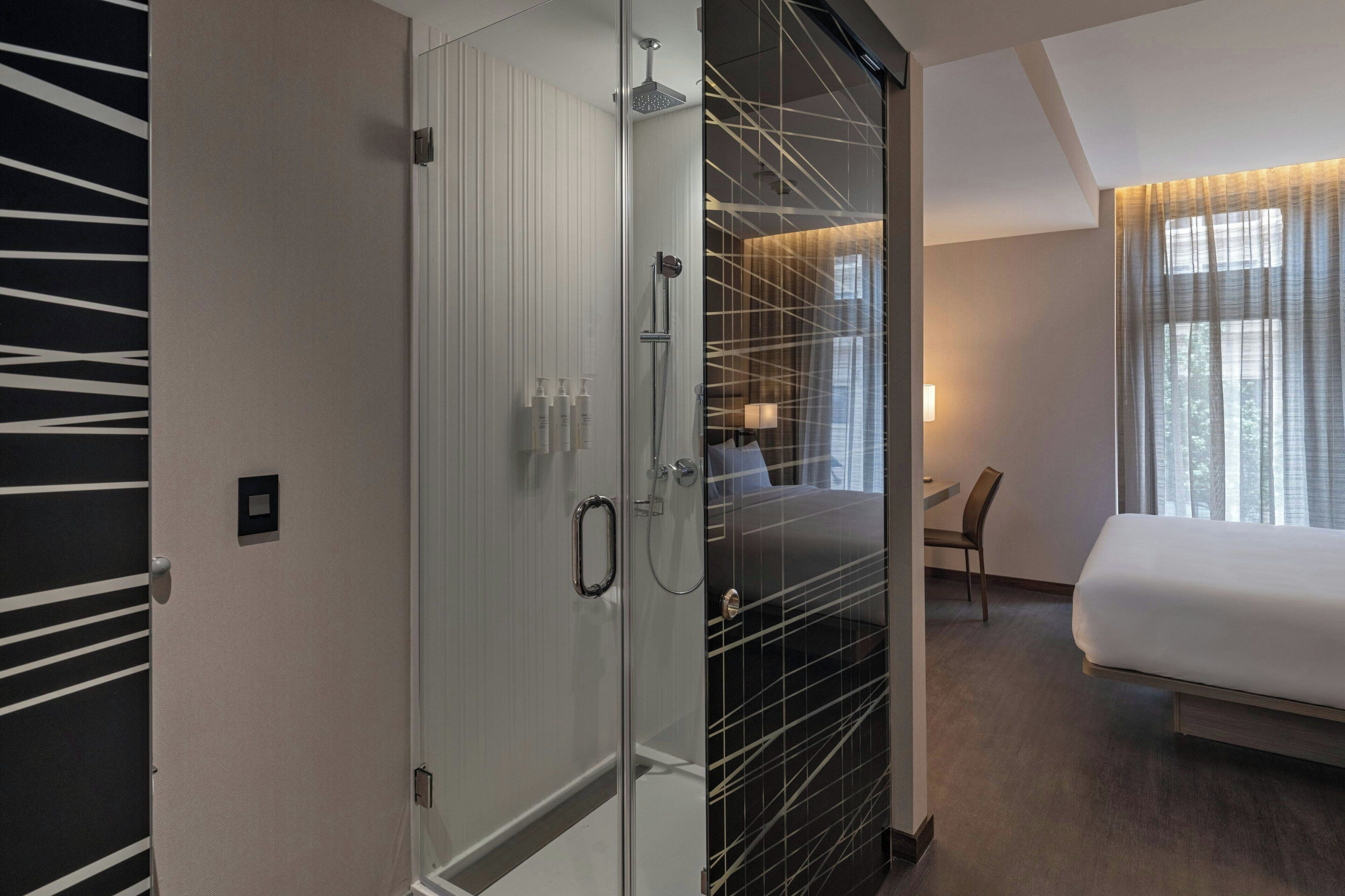 A modern hotel room with a glass shower enclosure, a bed, a desk, and a chair.