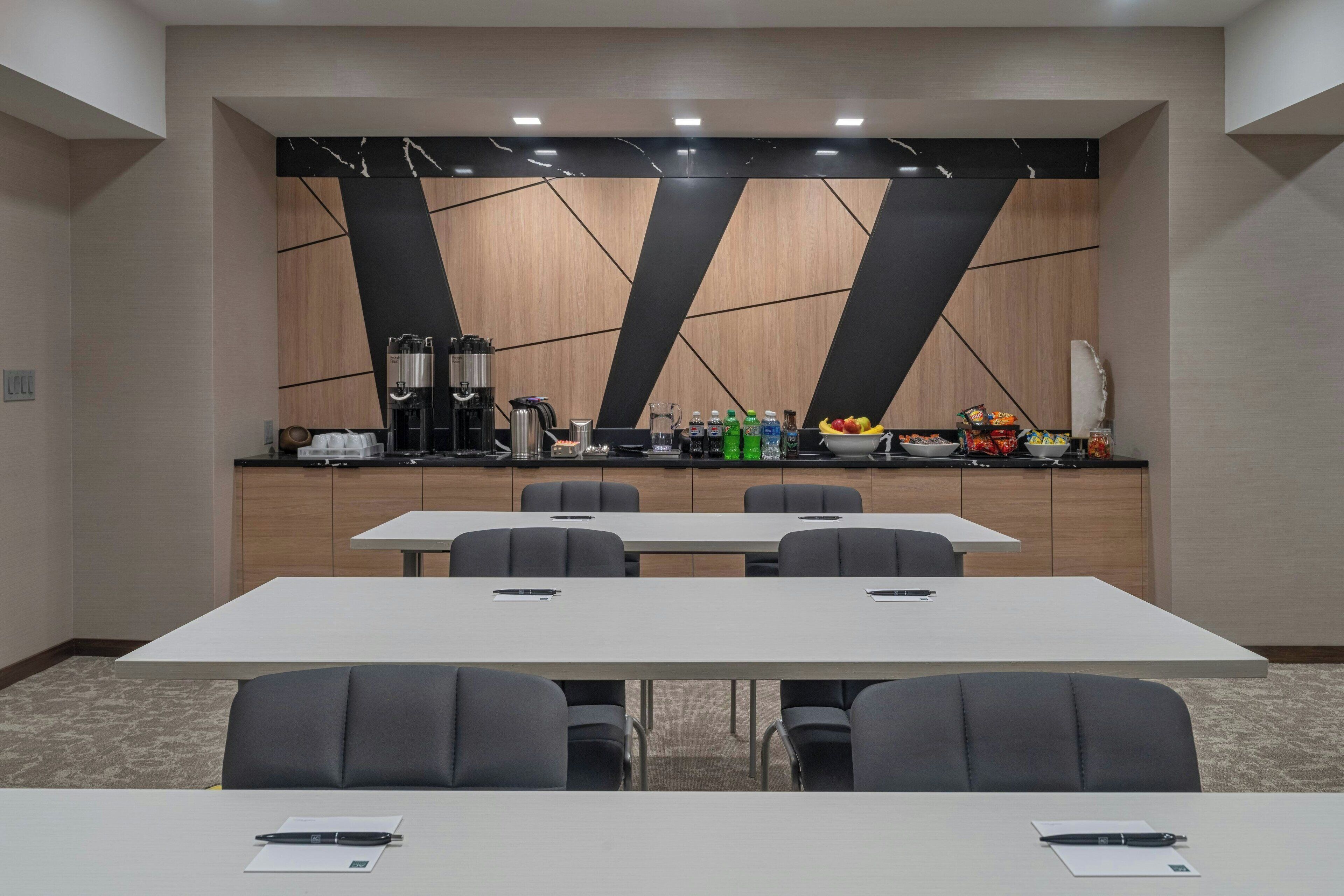 Une salle de conférence dotée d’une longue table et de chaises, d’un coin cuisine équipé d’appareils électroménagers et de boissons, et d’un décor mural moderne aux motifs géométriques.