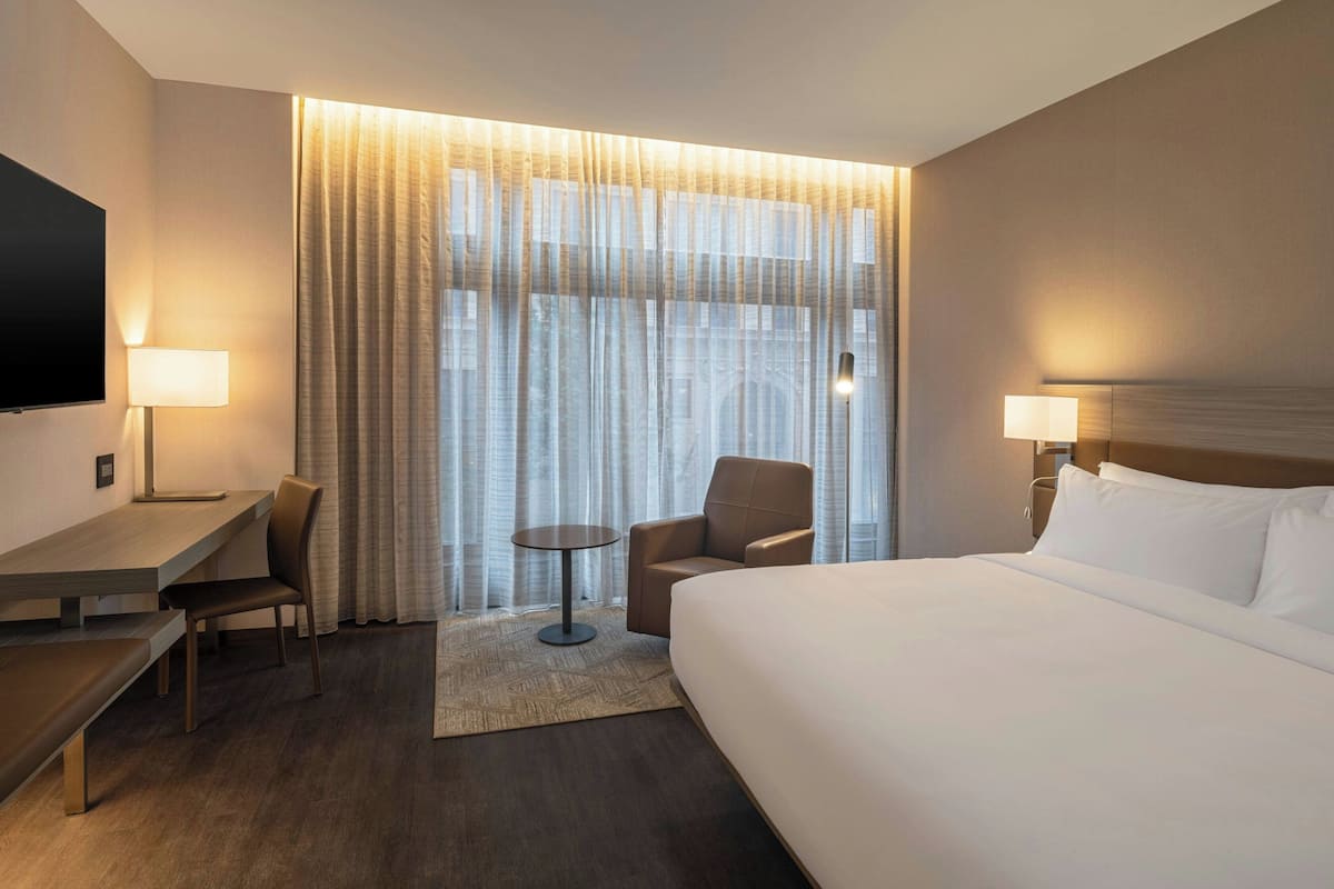 Habitación de hotel con una cama grande, un escritorio, una silla, un televisor y una ventana con cortinas.