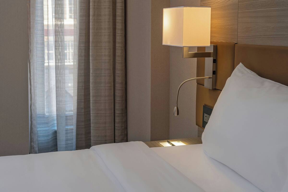 Una habitación de hotel con una cama bien hecha, una lámpara de pared y un ventanal con cortinas.