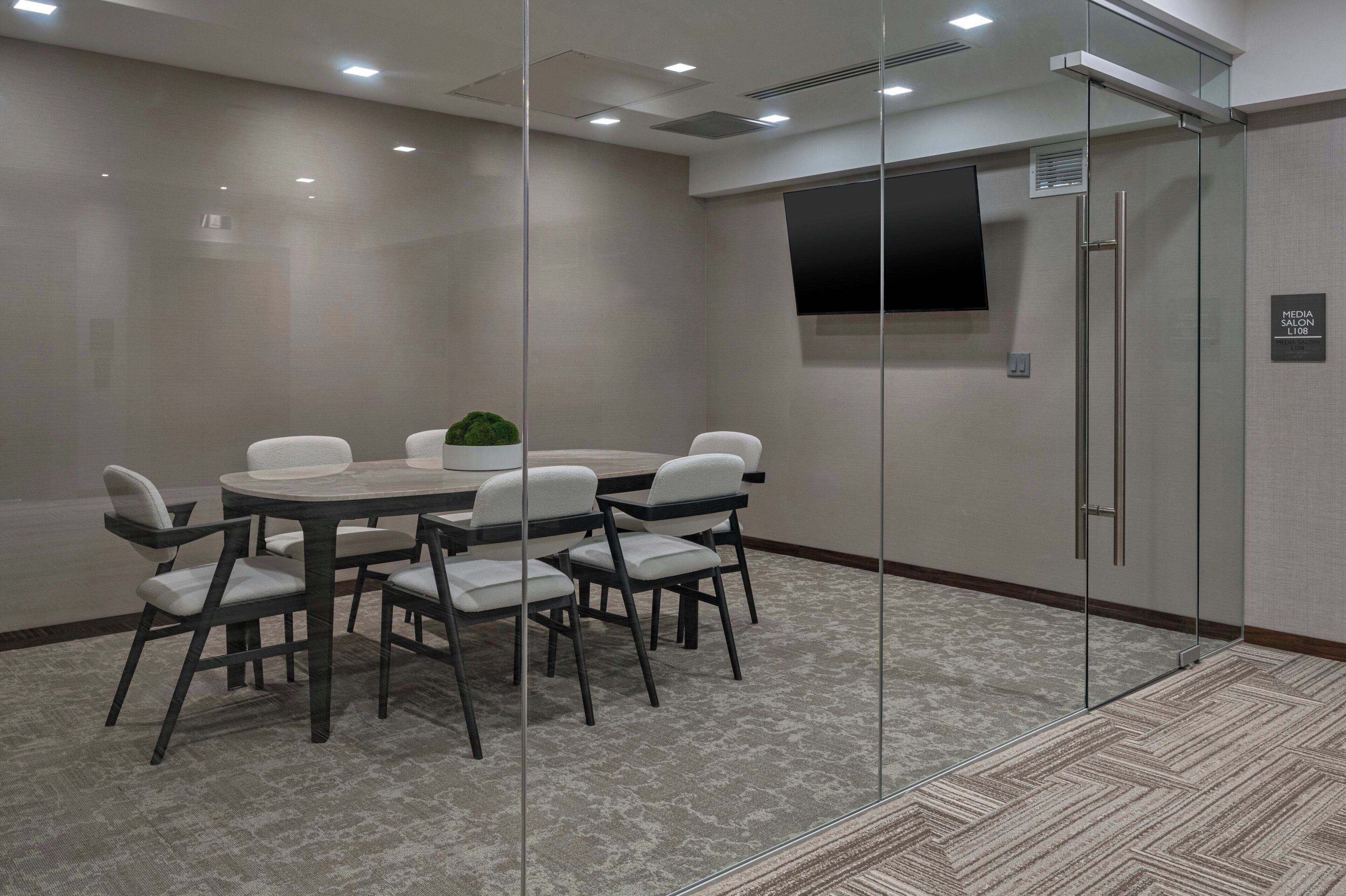 A conference room with a glass partition, a round table with chairs, a small plant, and a mounted TV on the wall.