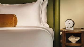 A neatly made bed with white linens and a brown throw pillow, a bedside table with a clock, and a framed abstract artwork on the wall.